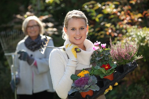 A young woman outdoors in a garden, holding a black tray filled with colourful flowering plants and shrubs, with a smile on her face. Behind her, an older woman with glasses, wearing a white jacket and scarf, is slightly out of focus, holding garden tools such as pruning shears and a rake. The garden features lush green foliage, flowering plants, and trees with red and orange leaves, suggesting a well-maintained landscaped outdoor space. The scene is illuminated by natural sunlight, creating a bright and vibrant atmosphere. The garden's layout includes a mix of lawn areas, flower beds, and paved pathways, typical of residential gardens in Purley, which benefits from professional gardening services like lawn mowing and plant care provided by Lawn Mowing Purley, specializing in outdoor maintenance and landscaping to enhance the natural beauty of local gardens.